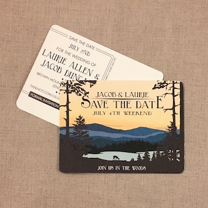 May include: Save the date card with a rustic woodland theme. The card features a sunset scene with mountains, trees, and a lake. The text reads "Jacob & Laurie Save the Date July 4th Weekend Join us in the Woods".