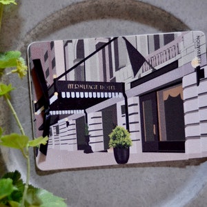 May include: A rectangular card featuring an illustration of the Hermitage Hotel with the word "RESERVATION" printed on the top right corner. The hotel's facade is depicted in shades of gray, black, and white, with a black awning and a small potted plant.