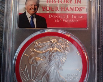 May include: A silver American Eagle coin with a red border, featuring the image of Lady Liberty, the year 2018, and the inscription "In God We Trust". The coin is encased in a clear plastic holder with a red label that reads "History in Your Hands", "Donald J. Trump", and "45th President".
