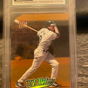 May include: A graded baseball card featuring a baseball player in a white uniform with a gold background. The card is graded 9.5 by a company called Premier Grading. The card features a photo of B.J. Upton, a shortstop for the Devil Rays. The card is from the Fleer Ultra series.