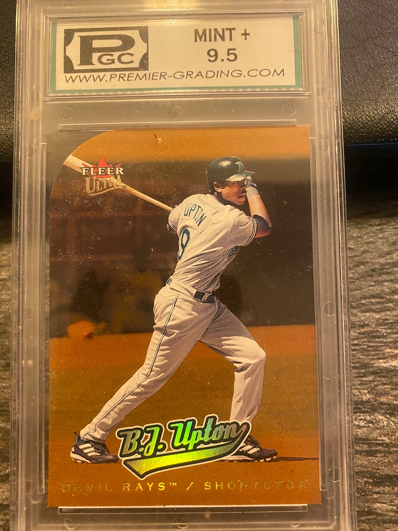 May include: A graded baseball card featuring a baseball player in a white uniform with a gold background. The card is graded 9.5 by a company called Premier Grading. The card features a photo of B.J. Upton, a shortstop for the Devil Rays. The card is from the Fleer Ultra series.