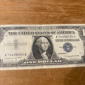 May include: A one dollar silver certificate from 1957, featuring a portrait of George Washington. The serial number is A 74498094 A. The text "THE UNITED STATES OF AMERICA" is printed in bold at the top of the bill.