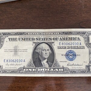 May include: A United States one-dollar silver certificate from 1957. The bill features a portrait of George Washington and the text "ONE DOLLAR" prominently displayed. The bill is green and black with intricate border designs.