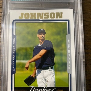 May include: A baseball card featuring Randy Johnson, a pitcher for the New York Yankees. The card is graded 9.5 by a professional grading company. The card features a photo of Johnson pitching, wearing a blue and white Yankees uniform. The card is from the 2005 Topps series.