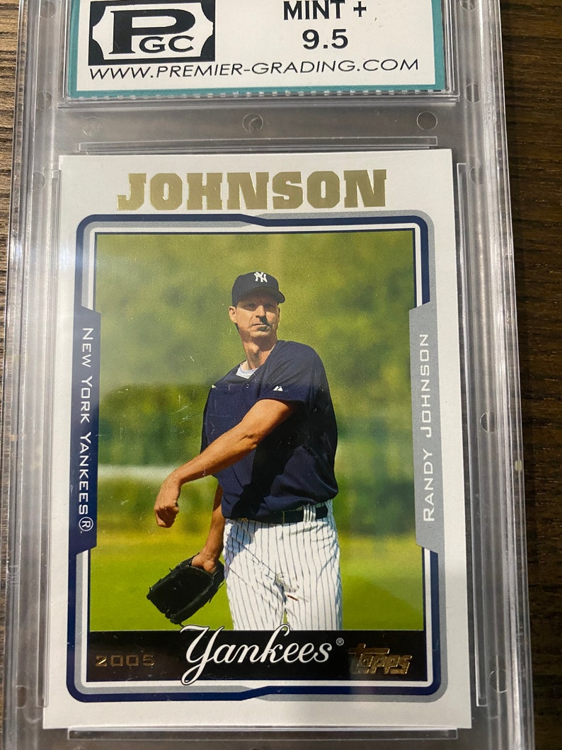 May include: A baseball card featuring Randy Johnson, a pitcher for the New York Yankees. The card is graded 9.5 by a professional grading company. The card features a photo of Johnson pitching, wearing a blue and white Yankees uniform. The card is from the 2005 Topps series.