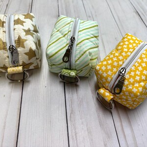 May include: Three small, square fabric pouches with zippers and key rings. One is cream with gold stars, another is mint green with stripes, and the third is yellow with white floral patterns. The pouches are likely for small items.