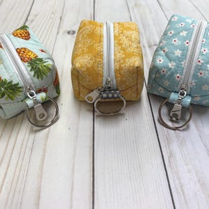 May include: Three small, square fabric pouches with silver zippers and key rings. One is light blue with pineapple print, another is yellow with a paisley pattern, and the third is light blue with white floral print.