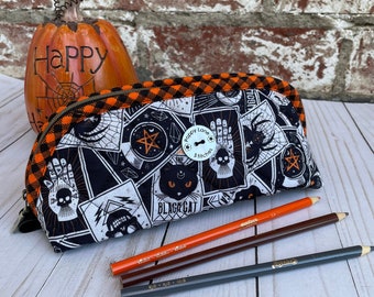 May include: A quilted fabric pencil case featuring a black and white pattern of tarot cards, skulls, and a black cat, accented with an orange and black checkered trim. A white circular button with the text Poppy Lane Stitches is attached.