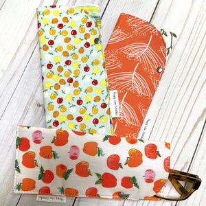May include: A collection of fabric eyeglass cases with various fruit and botanical patterns. One case features a light blue background with yellow, orange, and red fruit. Another has an orange background with white leaf designs. The third case has a white background with orange and pink fruit.