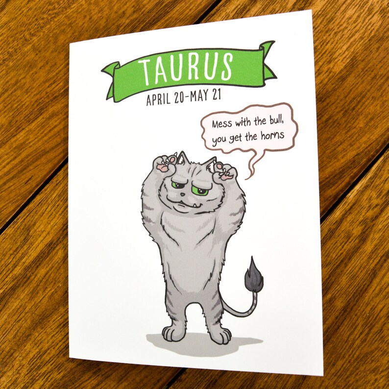 May include: A humorous illustration of a gray cat with green eyes, standing on its hind legs with its paws raised, resembling a bull. The text reads "TAURUS APRIL 20-MAY 21 Mess with the bull, you get the horns."