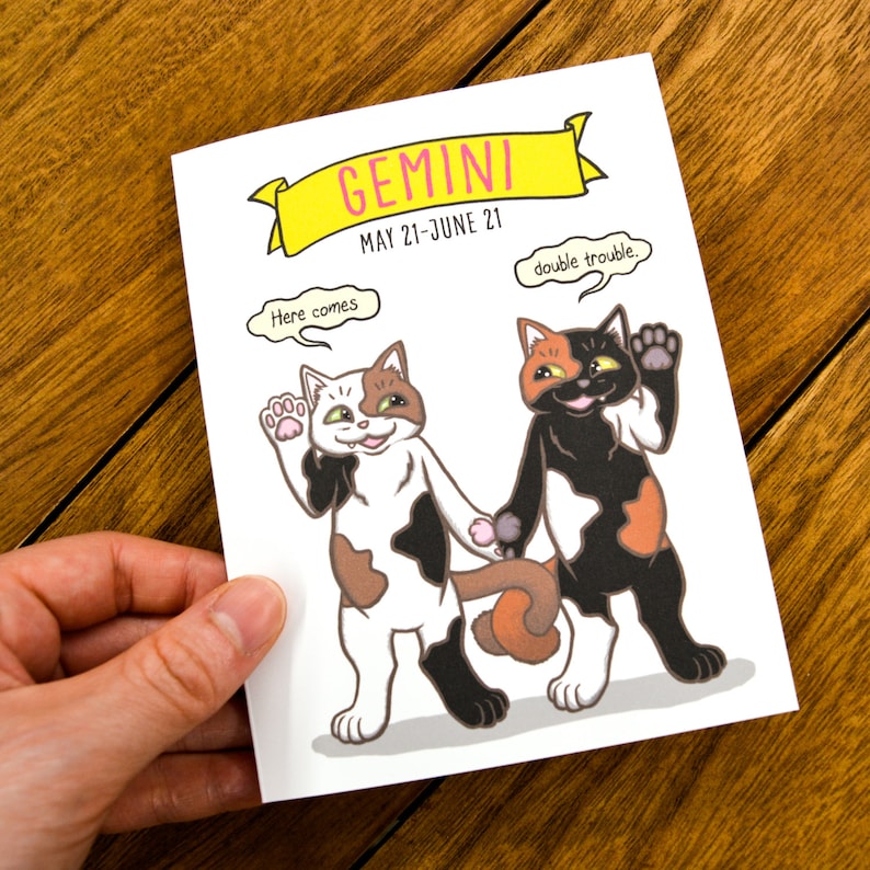 May include: A white card with a yellow banner that says "GEMINI MAY 21-JUNE 21". Two cartoon calico cats are holding hands. One cat has a speech bubble that says "Here comes" and the other cat has a speech bubble that says "double trouble."