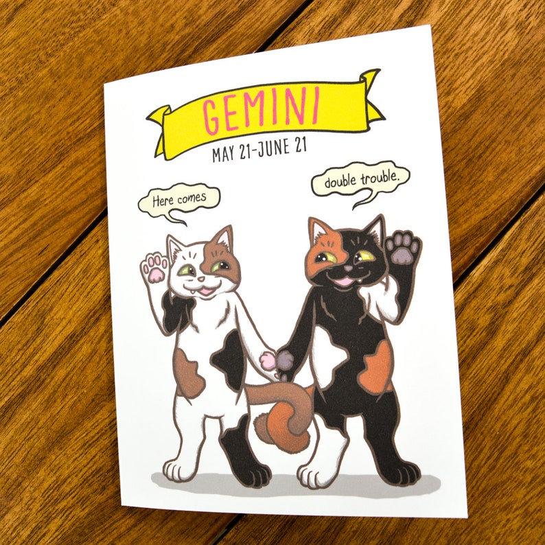 May include: A greeting card with a cartoon illustration of two calico cats. The card is white with a yellow banner at the top that says "GEMINI MAY 21-JUNE 21". One cat is saying "Here comes" and the other is saying "double trouble."