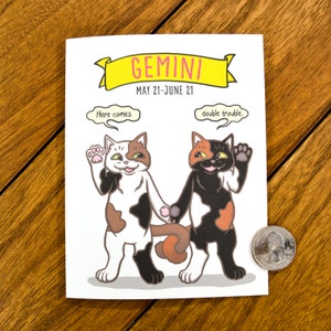 May include: A greeting card with a cartoon illustration of two calico cats holding hands. The card is white with a yellow banner at the top that says "GEMINI MAY 21-JUNE 21". One cat has a speech bubble that says "Here comes" and the other cat has a speech bubble that says "double trouble."