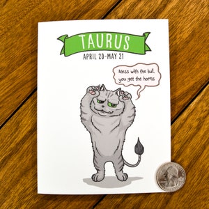 May include: A white card with a green banner that says "TAURUS" and "APRIL 20-MAY 21". A cartoon gray cat with a grumpy expression is standing on its hind legs with its paws raised. The cat has a speech bubble that says "Mess with the bull, you get the horns."