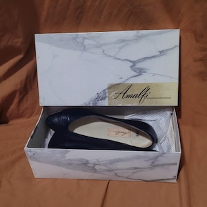 May include: A pair of navy blue Amalfi flats in a marble-patterned box. The box lid features the Amalfi logo in gold lettering. The shoes are displayed on white tissue paper inside the box.