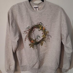 May include: A light gray sweatshirt with a crew neck and long sleeves. The front features a detailed wreath design with birds, berries, and foliage. The brand name "Jerzees" is visible on the collar.