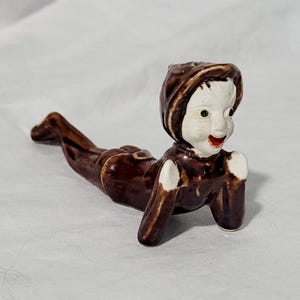 May include: A ceramic figurine of a person in a prone position, with a brown body and a white face. The figure is wearing a brown hood and has green eyes and a red mouth. The figurine is lying down on a white surface.