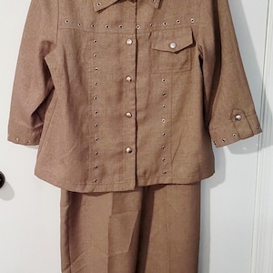 May include: A two-piece tan suit featuring a collared jacket with three-quarter sleeves, a button-up front, and decorative metal eyelets. The matching pants are straight-legged. The suit is made of a textured fabric.