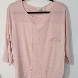 May include: A light pink pullover top featuring a round neckline, three-quarter length sleeves, and a single chest pocket. The garment is constructed with a sheer panel across the upper chest and shoulders, finished with a banded hem at the waist.