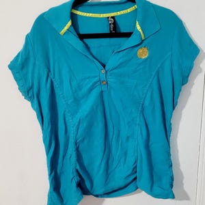 May include: A turquoise polo shirt with short sleeves, a collared neckline, and a three-button placket. The collar and inner neckline feature a bright yellow trim. A gold emblem is on the left chest. The shirt is hanging on a black hanger.
