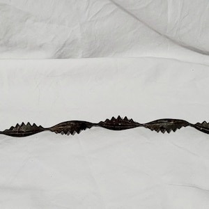 May include: A dark metal, wavy-shaped tool with jagged edges. The tool has a hook on one end and a leaf-like shape on the other. The object is set against a white, wrinkled background.