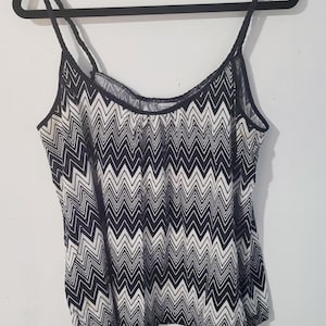 May include: A black and white chevron patterned camisole top with thin spaghetti straps. The top is hanging on a black hanger against a white wall. The fabric has a repeating zigzag design.