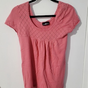 May include: A short-sleeved, coral-colored top with a scoop neckline and diamond-patterned detailing. The top has a gathered waist and is hanging on a black hanger. The tag reads "It's Our Time".