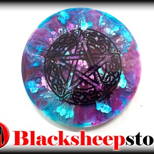 Pode incluir: Um coaster redondo de resina com um design de pentagrama preto cercado por um padrão de nó celta. O fundo é uma mistura de azul, roxo e branco.