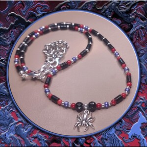 May include: A necklace with a silver spider pendant, featuring black, red, and blue beads. The pendant is a detailed silver spider. The necklace is displayed on a tan surface with a blue and red abstract background.