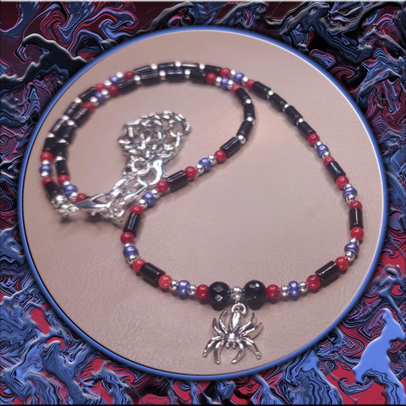 May include: A necklace with a silver spider pendant, featuring black, red, and blue beads. The pendant is a detailed silver spider. The necklace is displayed on a tan surface with a blue and red abstract background.