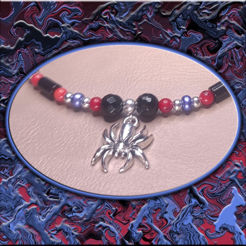 May include: A necklace with a silver spider charm, strung with black, red, silver, and purple beads. The background is an abstract design in blue and red, with the necklace in an oval frame.