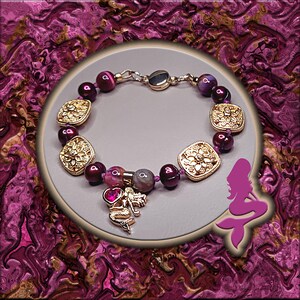 May include: A bracelet featuring gold-colored square beads with floral designs, interspersed with round, deep purple beads. A gold-colored charm of a dragon with a heart pendant hangs from the bracelet. The bracelet is set against a purple background.
