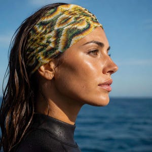 May include: A wide, stretchy fabric headband featuring a repeating pattern of yellow, green, and white sunflower-like petals. The headband is worn around the head, sitting across the forehead and covering the ears, made of a lightweight material.