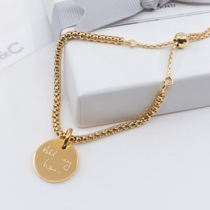Op de afbeelding: Verstelbare gouden armband met een ronde bedel met de tekst "Del my how." De armband ligt op een witte doos met een grijze strik.