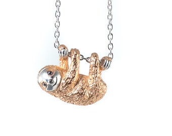 May include: A gold-toned sloth pendant hangs from a silver chain. The pendant is detailed with a textured surface and a smiling face.