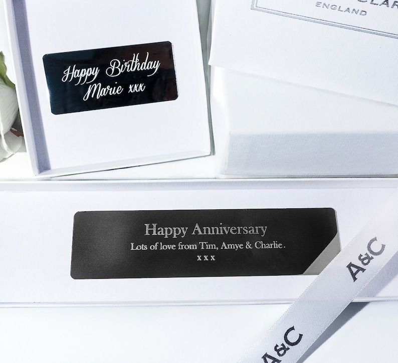 Op de afbeelding: Twee witte cadeaudozen met zwarte gegraveerde etiketten. De bovenste doos heeft de tekst "Happy Birthday Marie xxx". De onderste doos heeft de tekst "Happy Anniversary Lots of love from Tim, Amye & Charlie. XXX". Beide dozen hebben een wit lint met de tekst "A&C" erop gedrukt.