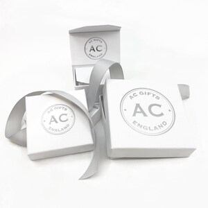 Op de afbeelding: Drie witte cadeaudozen met een zilveren logo met de tekst "AC Gifts England". De dozen zijn van verschillende grootte en zijn op elkaar gestapeld.
