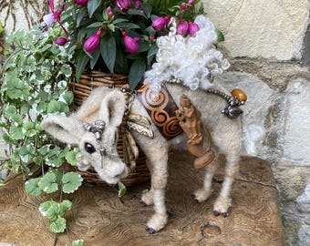 May include: A handcrafted, felted animal sculpture with a cream-colored body, adorned with decorative elements. It features a leather harness with a swirl design, a wooden carving, and silver-toned embellishments. The sculpture is posed on a wooden surface, with a basket of flowers in the background.