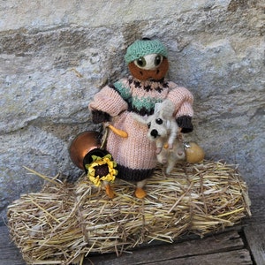May include: A knitted doll with a green hat and brown eyes is holding a small knitted animal. The doll is wearing a pink and brown sweater and is sitting on a pile of hay.