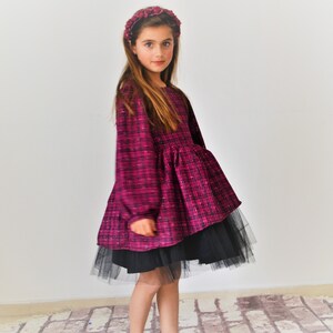 May include: A young girl wearing a pink and black plaid dress with a black tulle underskirt. She is wearing a burgundy flower headband and has long brown hair.