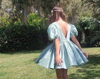 May include: A young person wearing a light blue dress with puffed sleeves and a deep V-neck. The dress is flowing and has a gathered skirt. The person is standing on a grassy lawn.
