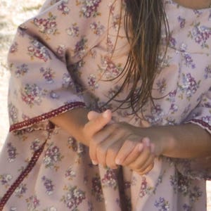May include: A young girl wearing a pink dress with a floral pattern. The dress has a ruffled hem and a burgundy trim around the sleeves.