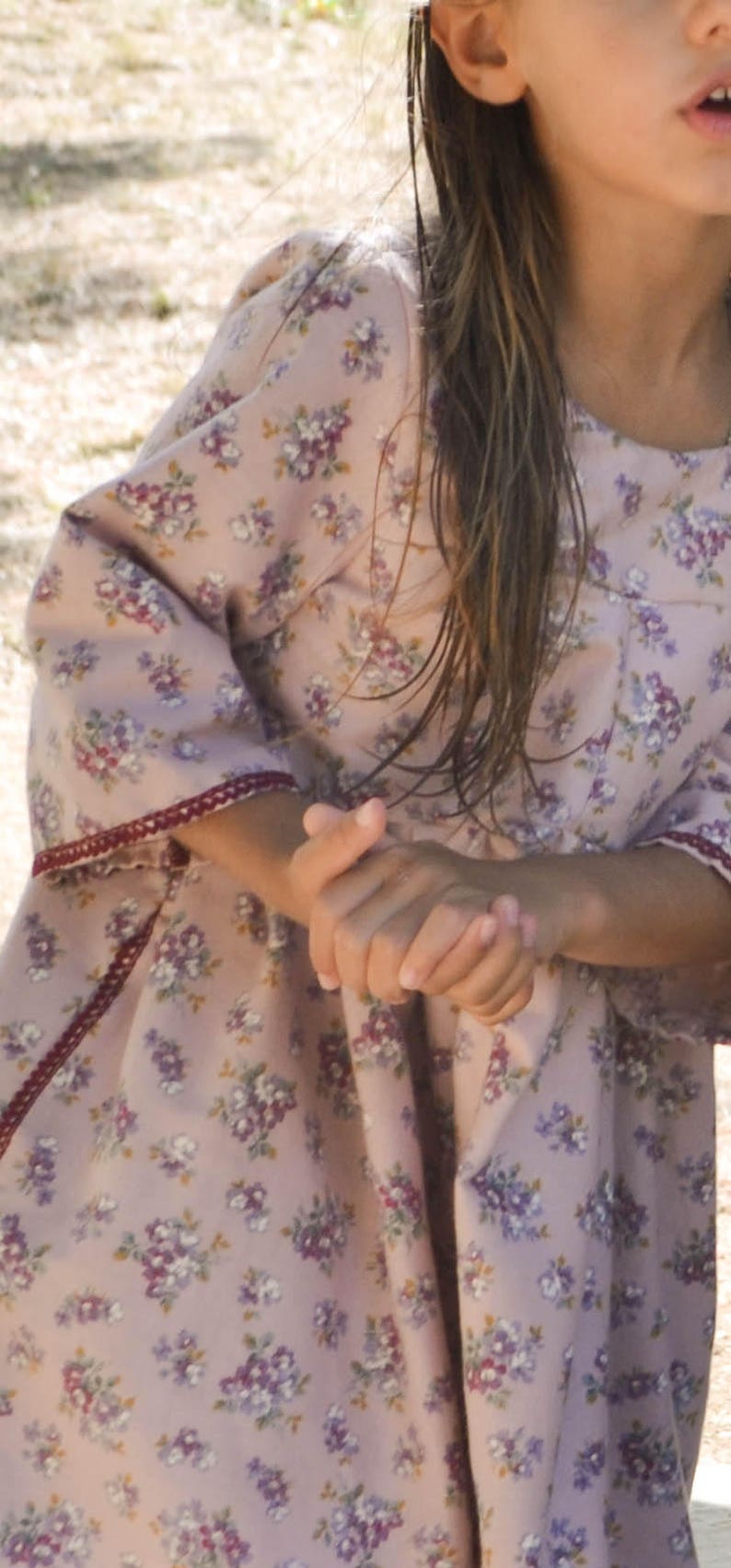May include: A young girl wearing a pink dress with a floral pattern. The dress has a ruffled hem and a burgundy trim around the sleeves.