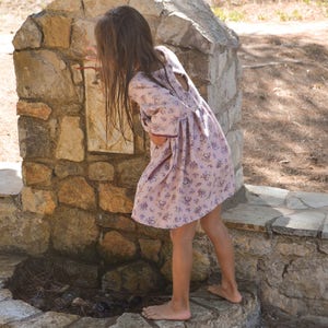 May include: A young girl in a floral dress with purple accents stands near a stone fountain. The fountain is made of stacked stones and has a small pool of water at the bottom.