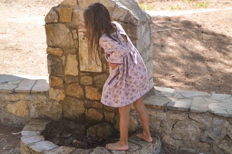 May include: A young girl in a floral dress with purple accents stands near a stone fountain. The fountain is made of stacked stones and has a small pool of water at the bottom.