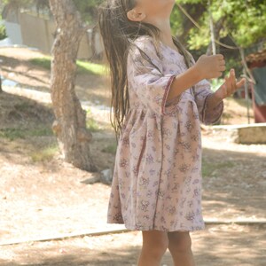 May include: A young girl wearing a pink floral dress with red trim stands in a grassy area and looks up at the sky.