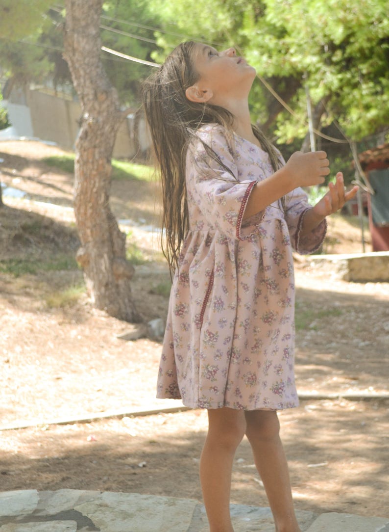 May include: A young girl wearing a pink floral dress with red trim stands in a grassy area and looks up at the sky.