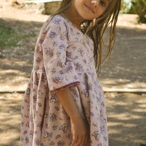 May include: A young girl wearing a pink and white floral dress with a burgundy trim. She has long brown hair and is looking at the camera.