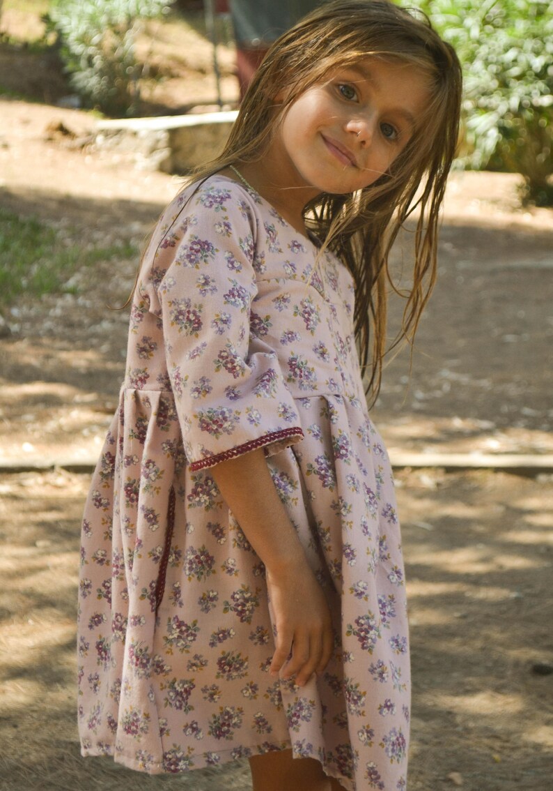 May include: A young girl wearing a pink and white floral dress with a burgundy trim. She has long brown hair and is looking at the camera.