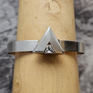 May include: A silver ring with a geometric triangular design. The ring features a polished band and a prominent triangle shape at the center, with a small, clear gemstone set within the triangle. The ring is displayed on a wooden ring holder.
